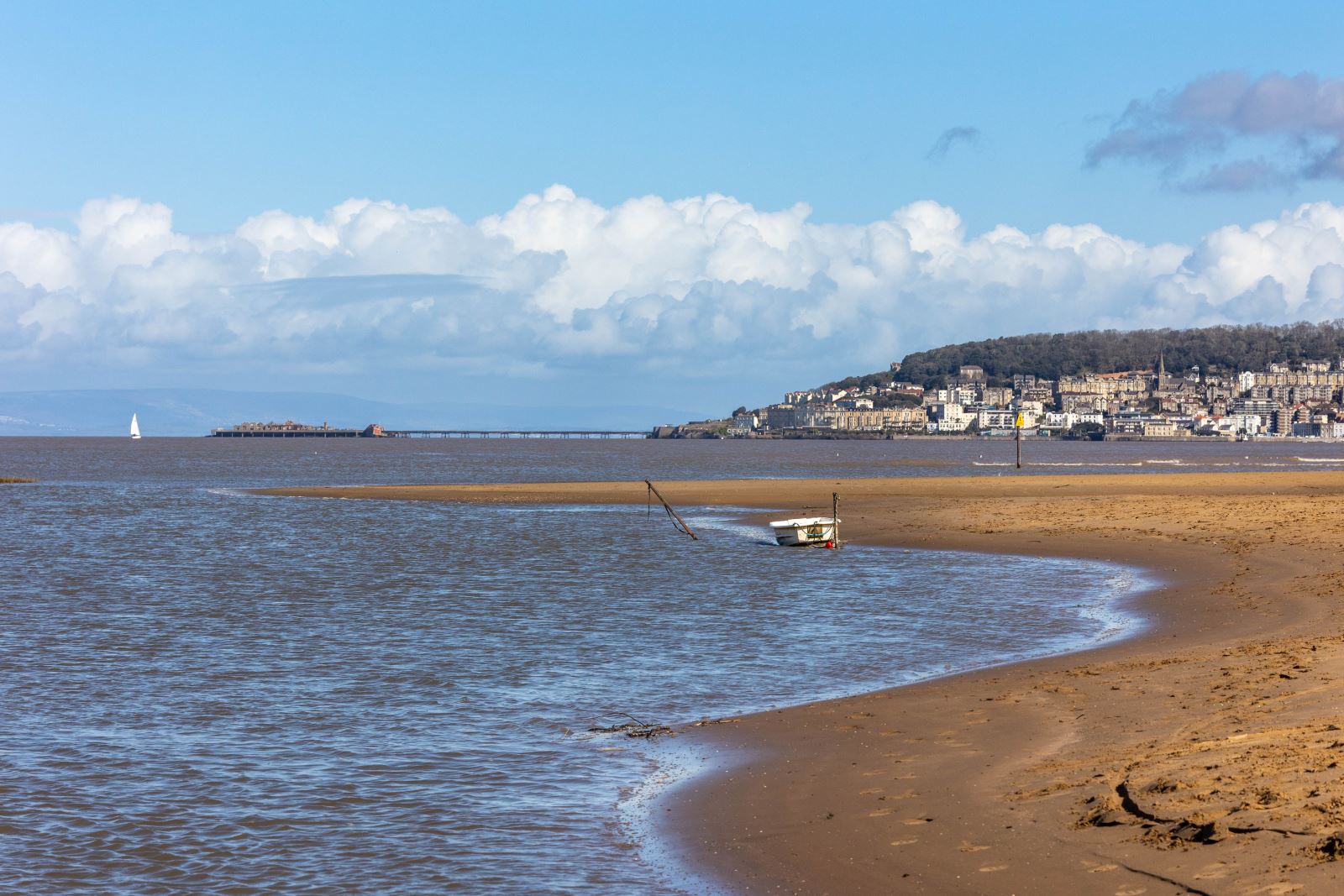 A seaside scene featuring a boat moored on the edge of the water on a sandy inlet with a town in the background